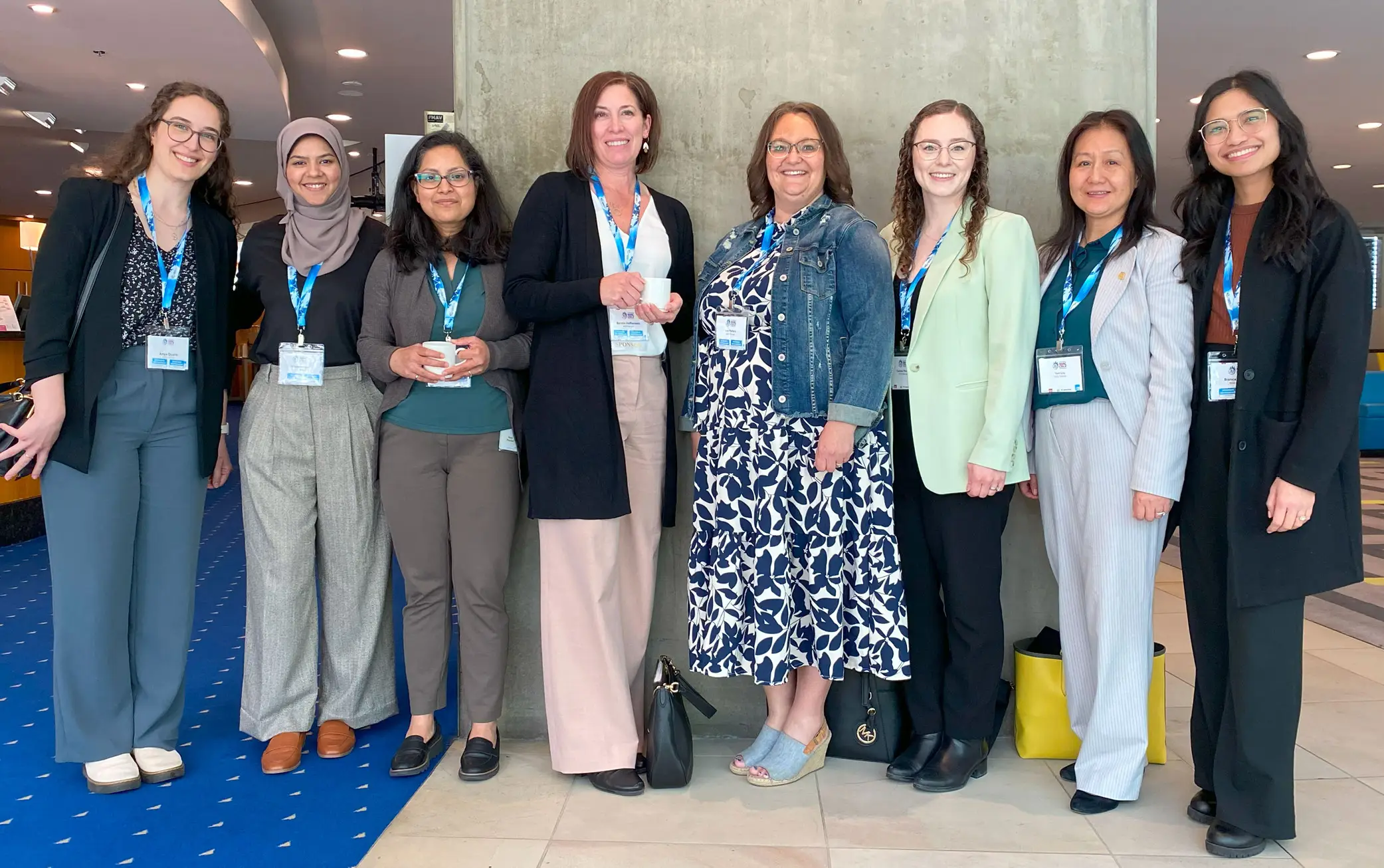 A group of eight KGS Group women employees, dressed in casual business attire and wearing name badges, smile together while posing at a conference event.