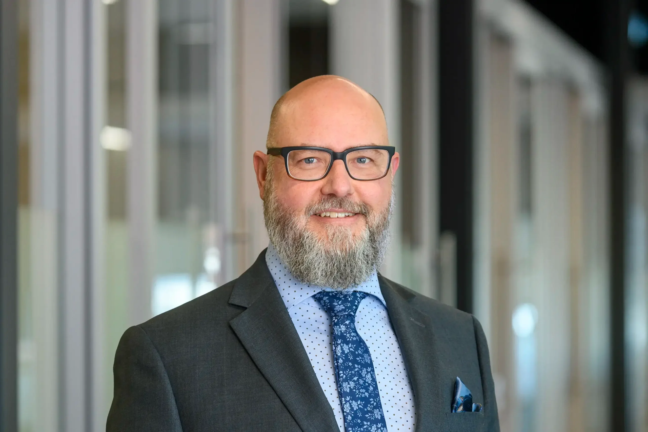 Ashley Dent stands confidently in a grey suit within a modern office setting.