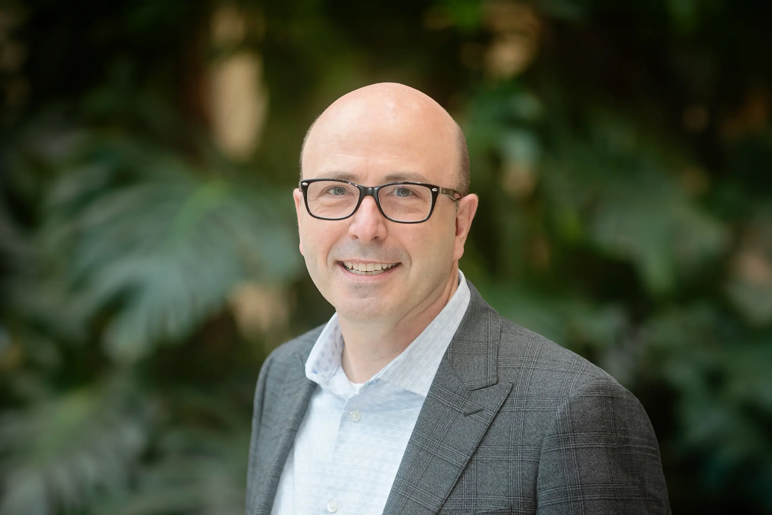 Bruce Belmore smiles warmly in his plaid suit while standing before a backdrop of green trees.
