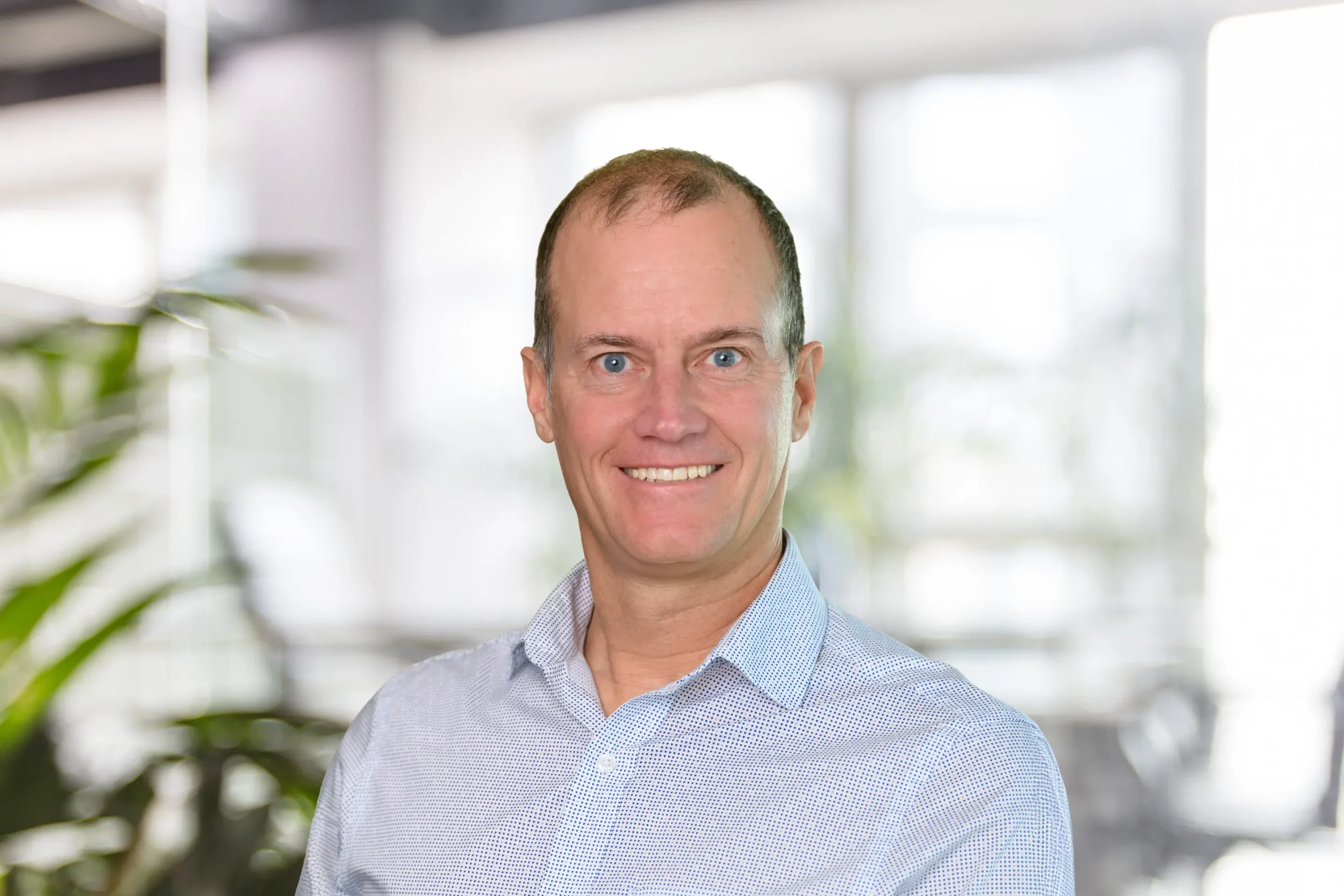 Craig Rowbotham poses in a light-blue button-down shirt, with a blurred office backdrop behind him.