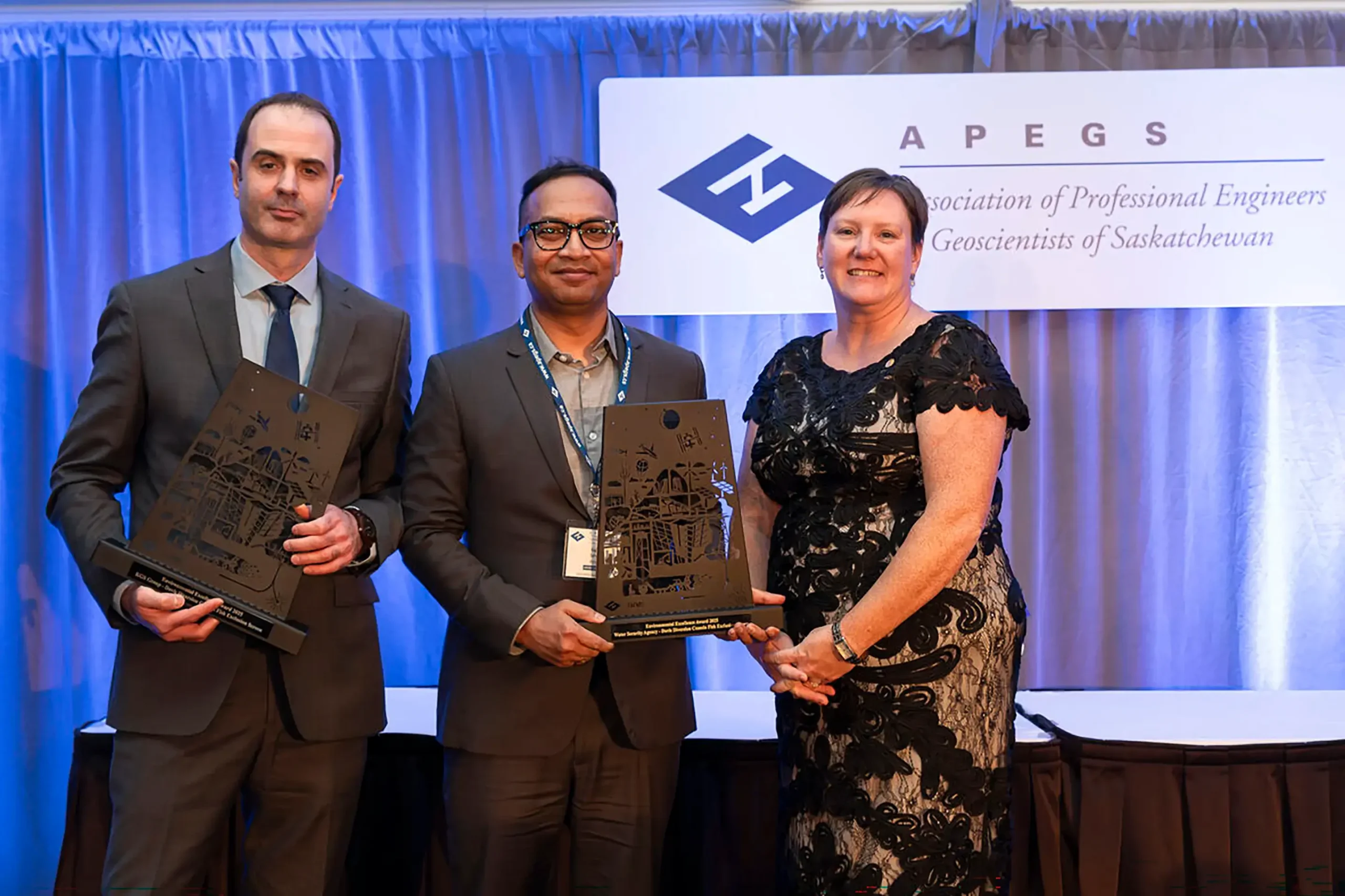 Two KGS Group employees in business attire accepting their Environmental Excellence Award from a representative of the APEGS.