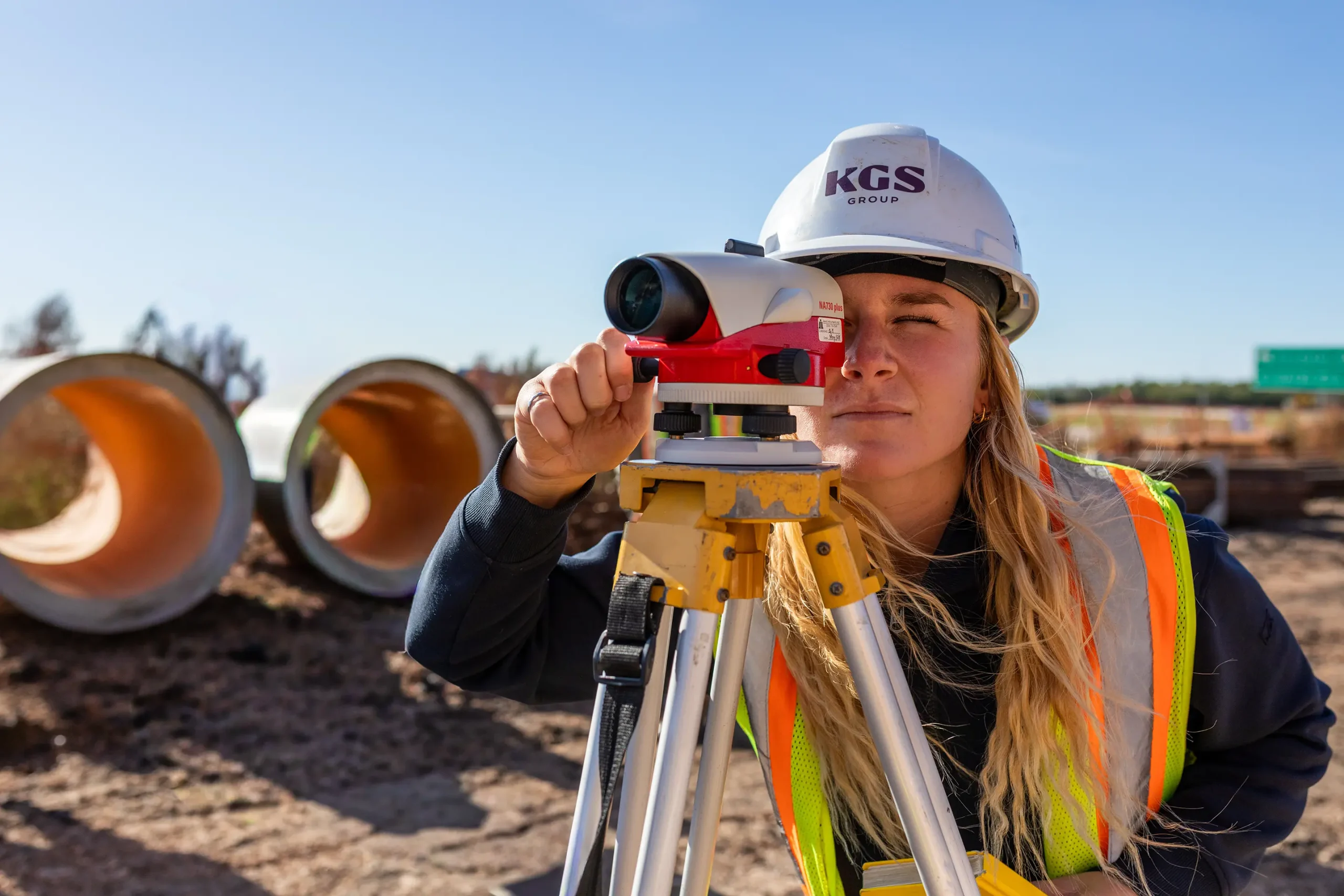 A woman wearing a hi‑visibility vest and hard hat peers into surveying equipment.