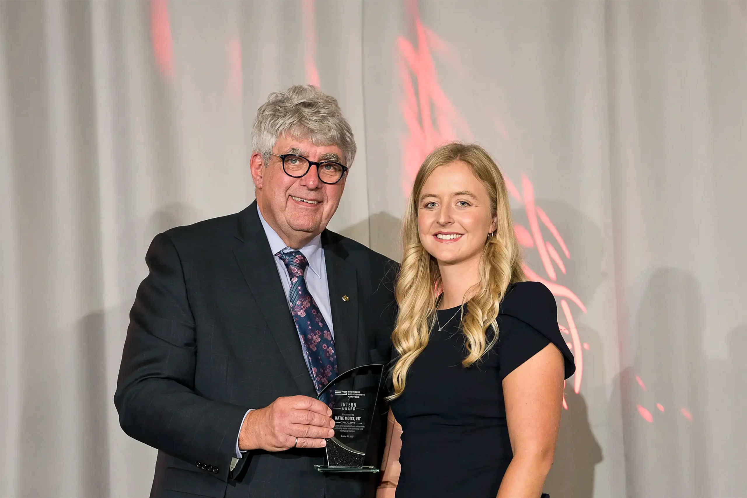 Katie Moist, dressed in a black gown, stands before a white curtain backdrop as she receives an award from a presenter wearing a black suit and tie.