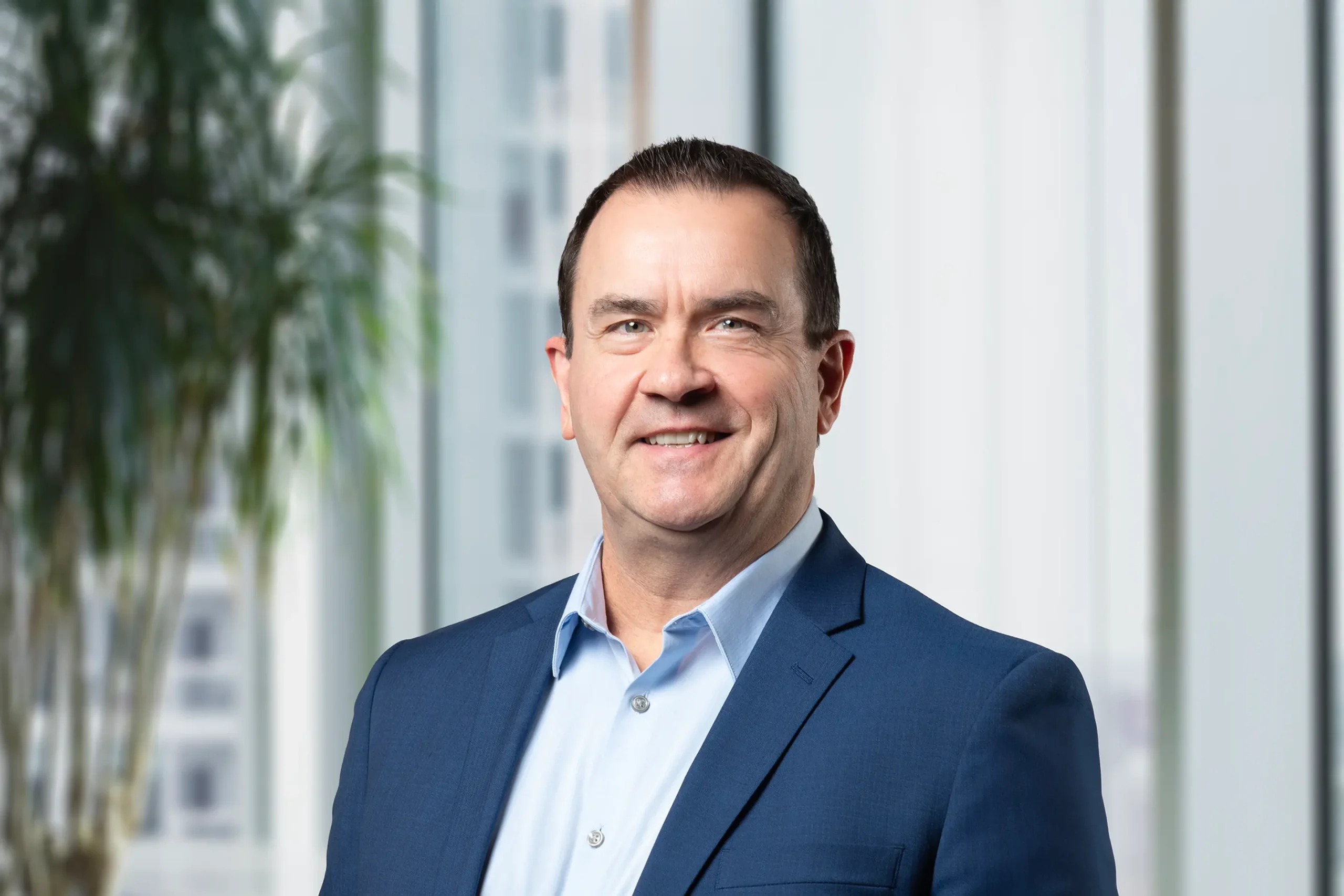 Peter Hassan stands confidently in a navy blue suit, framed by a modern office interior with a green plant.