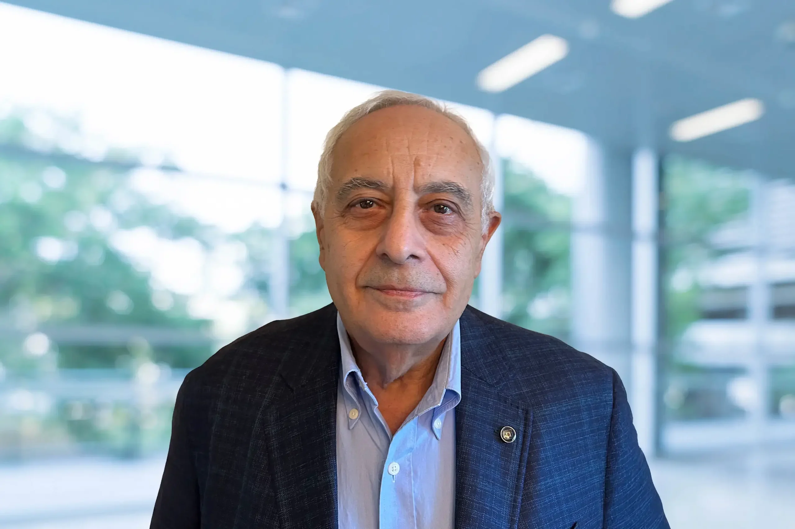 Albert Mikhail stands in a navy-blue suit jacket, offering a subtle smile as he poses in front of large glass windows that overlook a backdrop of trees.