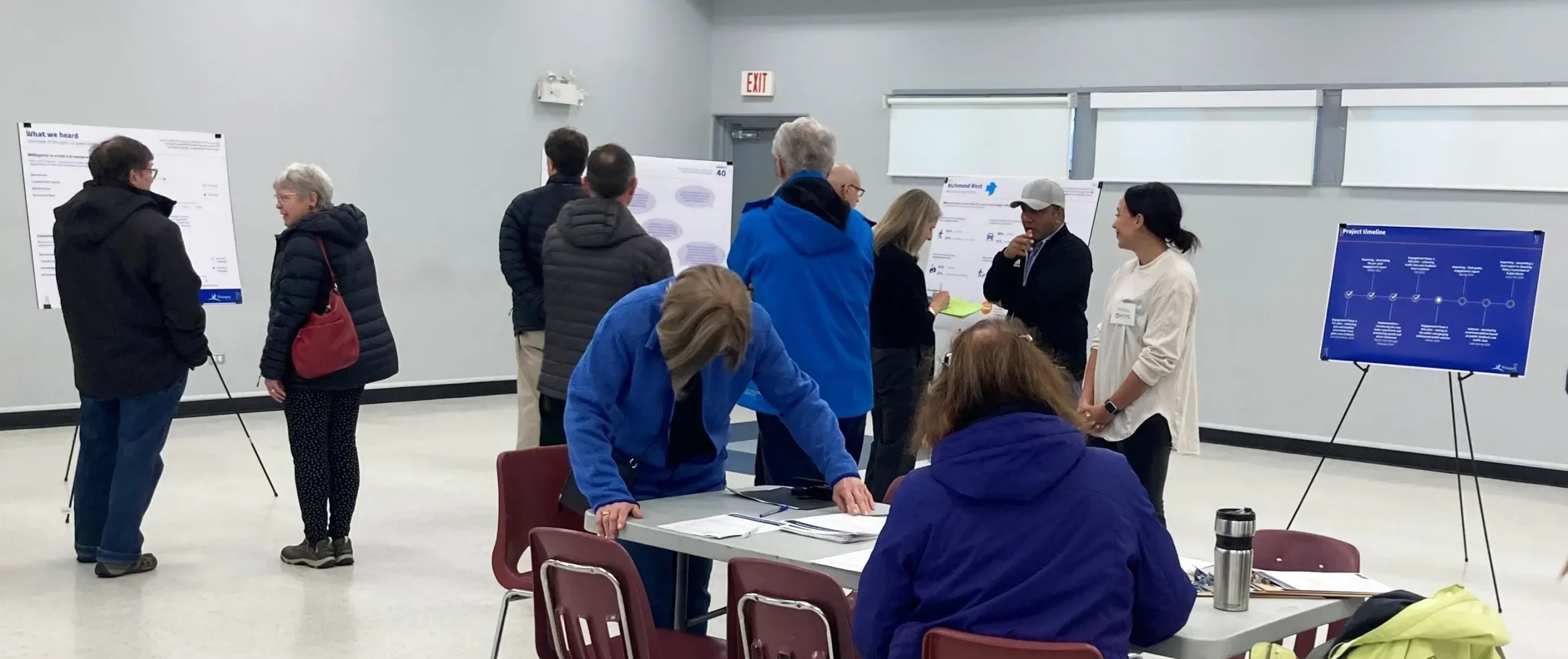 People at an indoor community meeting reviewing posters on easels and discussing at tables, with a project timeline display visible.