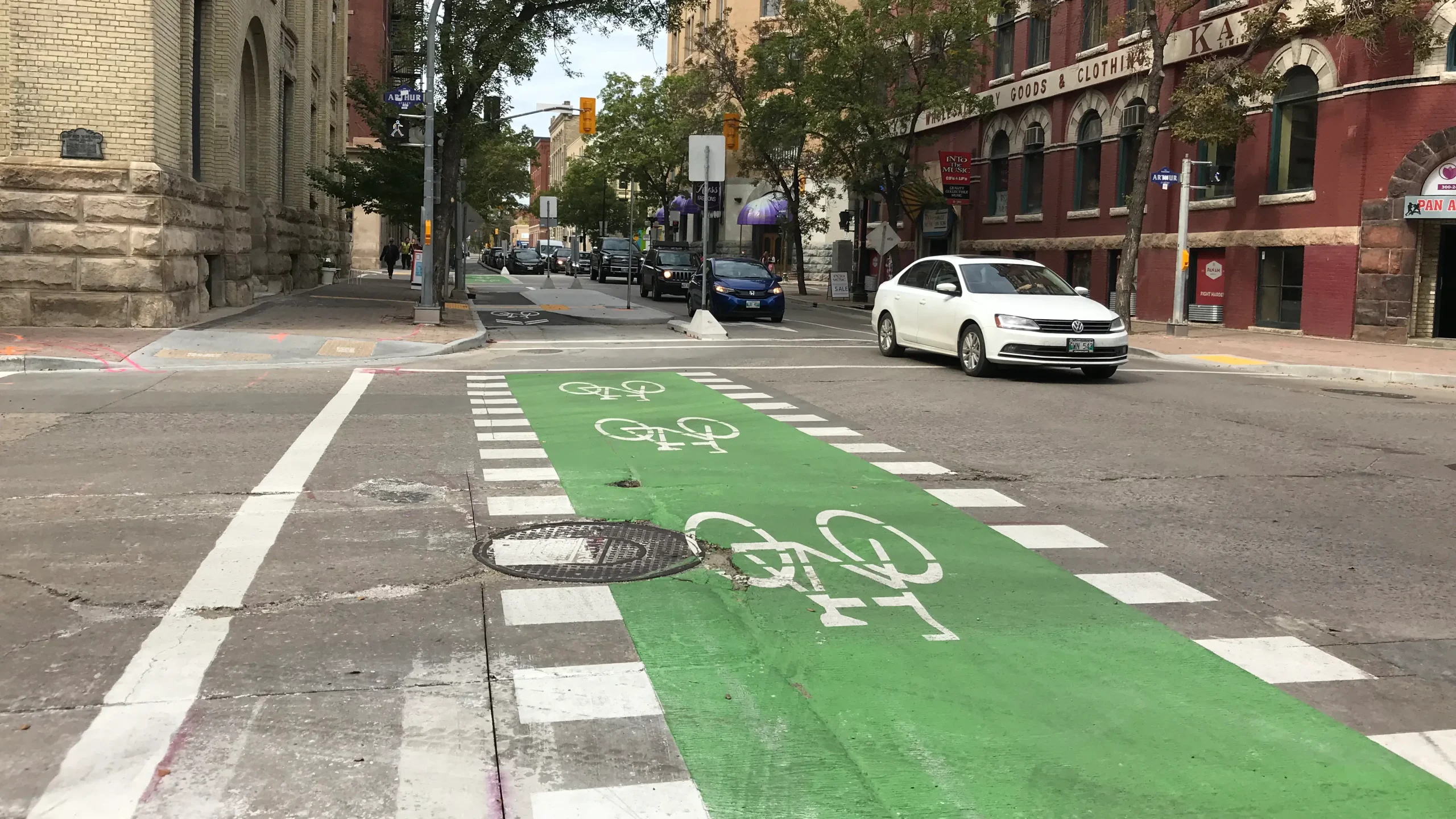 Urban intersection with a green bike crossing, marked by white bicycle symbols, with cars passing and buildings lining the street.