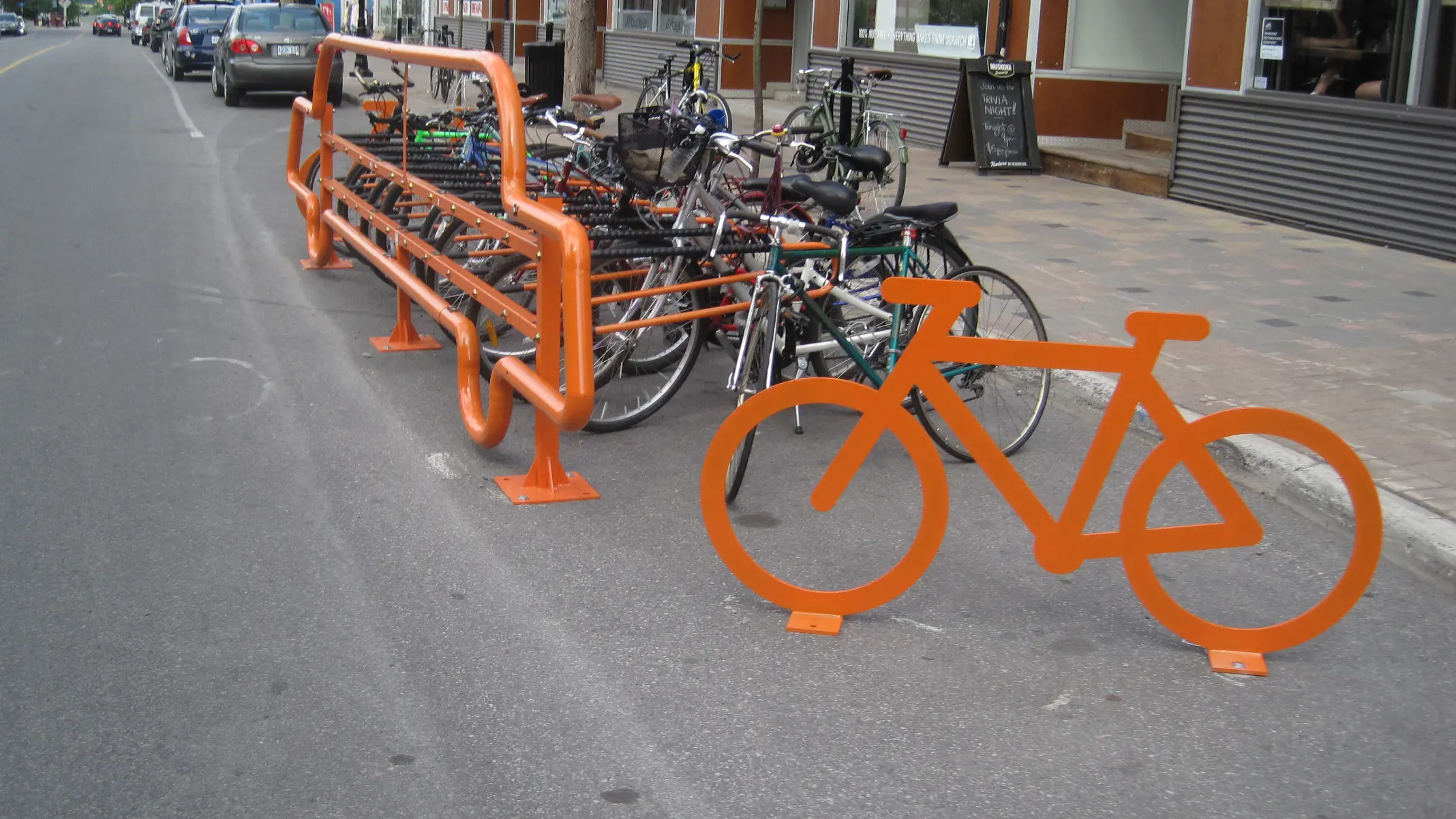 Orange bike racks filled with bicycles along a curbside lane, separated from traffic by barriers, with parked cars and storefronts nearby.