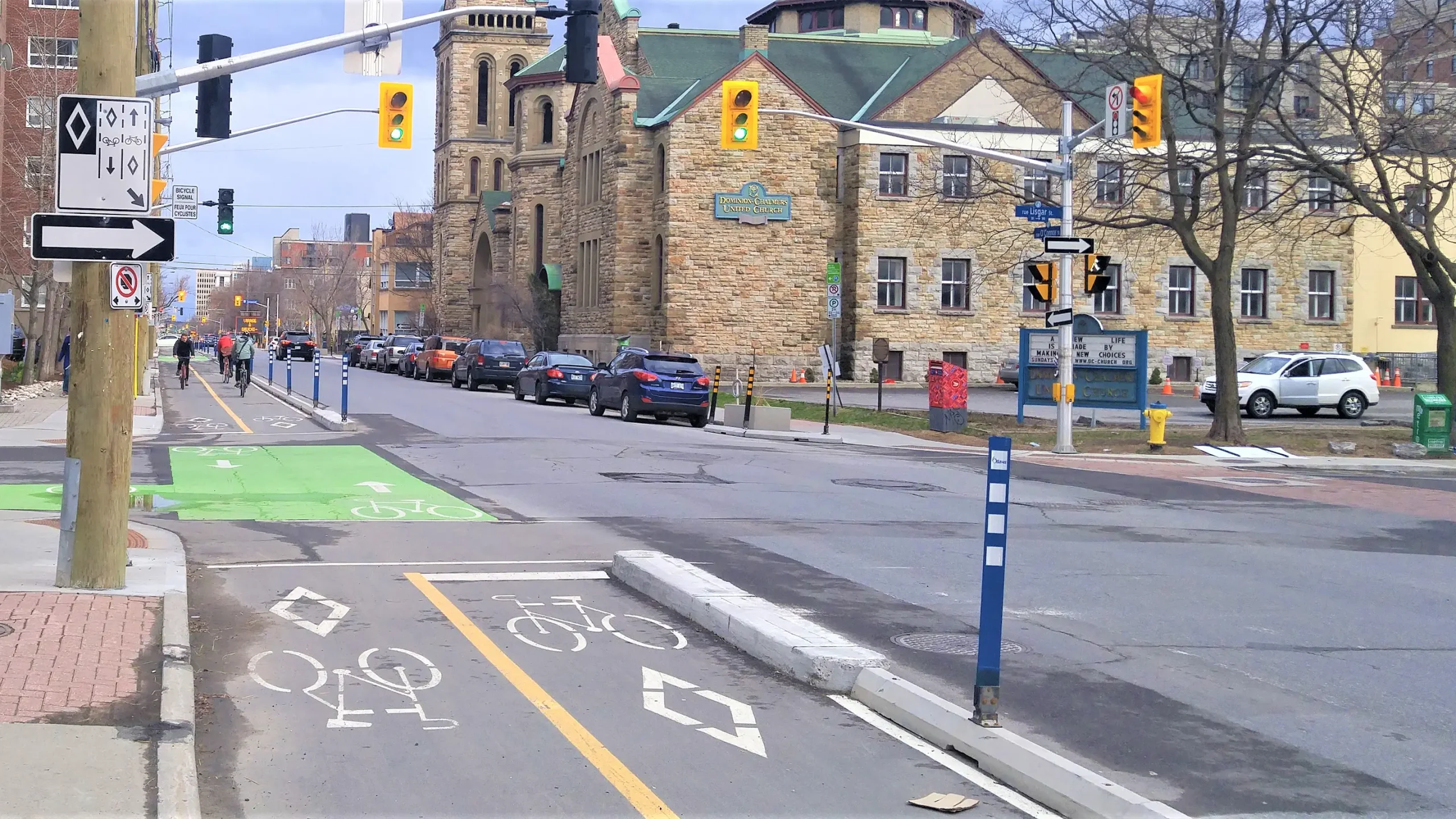 Intersection with a green protected bike lane, separated by posts, with cyclists riding and cars nearby.